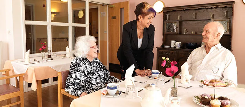 A carer serving afternoon tea to two residents in the dining room